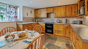 A kitchen with wooden cabinets, a stone tile floor, a dining table with chairs, and a window with floral curtains.