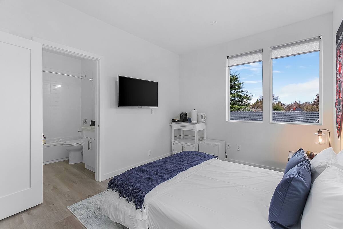 A modern bedroom with a large bed, a flat-screen TV mounted on the wall, a window with a view of trees, and a white wardrobe.