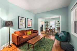 A living room with a sofa, a coffee table, a floor lamp, and a green armchair. There is a dining area with a table and chairs visible through an open doorway. - Charming 2 BR house in Old Hickory Village, a short walk from the lake road  (Nashville)