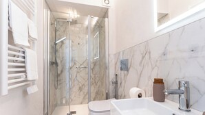 Uma casa de banho moderna com chuveiro em cabine de vidro, lavatório branco e parede revestida a mármore.