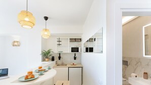 Cozinha moderna com armários brancos, micro-ondas embutido e pequena geladeira. Há uma mesa de café da manhã com croissants e suco no balcão.
