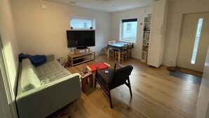 Een knus woongedeelte met een bank, een televisie op een houten dressoir, een eettafel met stoelen, een kleine keukenhoek en een deur naar buiten.