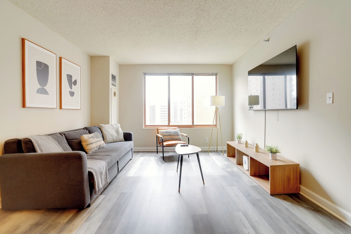 Una sala moderna con sofá, mesa de centro, sillón, televisor y ventana con vista a la ciudad.