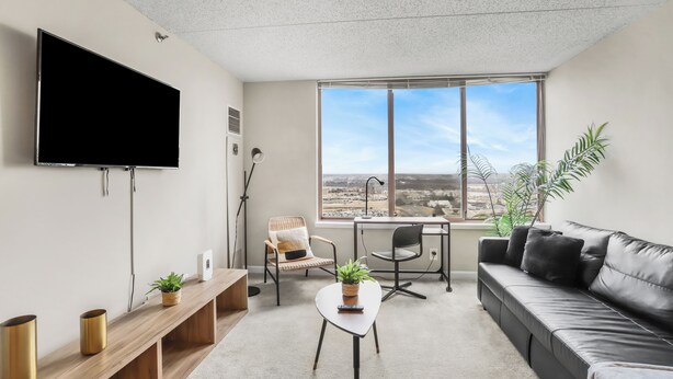 Ein modernes Wohnzimmer mit Flachbildfernseher, einer Couch, einem Schreibtisch und einem großen Fenster mit Blick auf die Stadt.