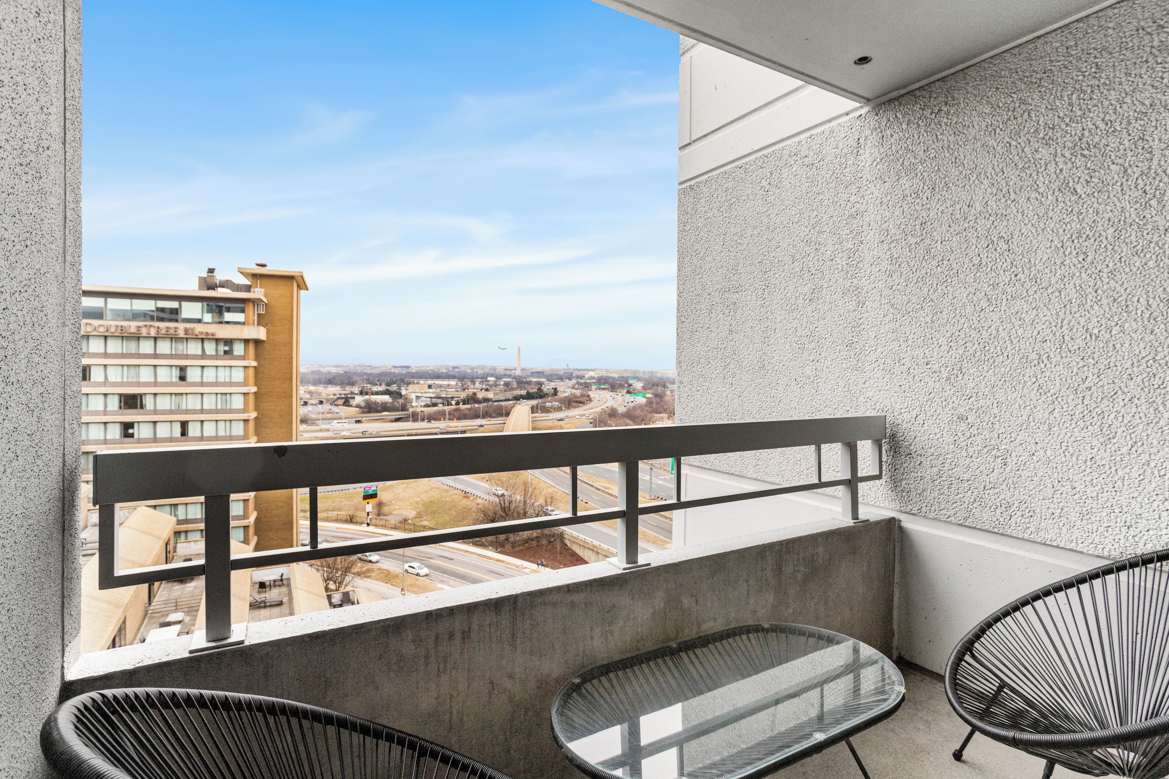 A balcony with a concrete floor, two black wire chairs, and a glass table. A building with a sign reading "DOUBLETREE" is visible in the background.