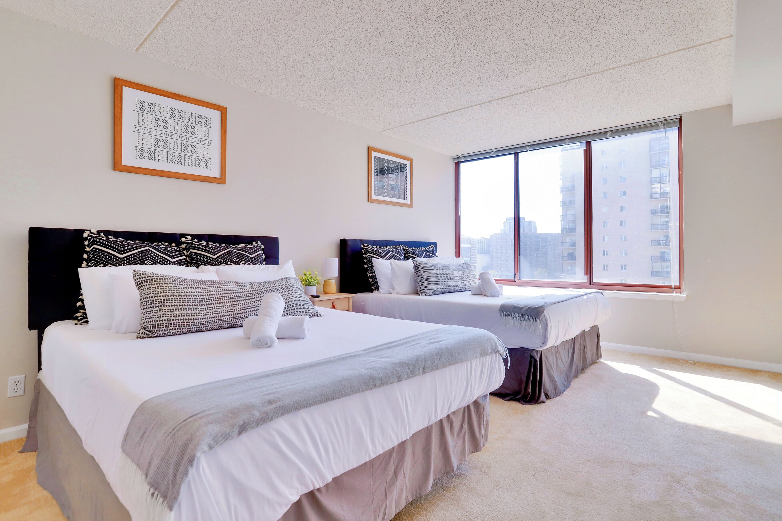 A hotel room with two beds, a large window with a city view, and a framed artwork on the wall.