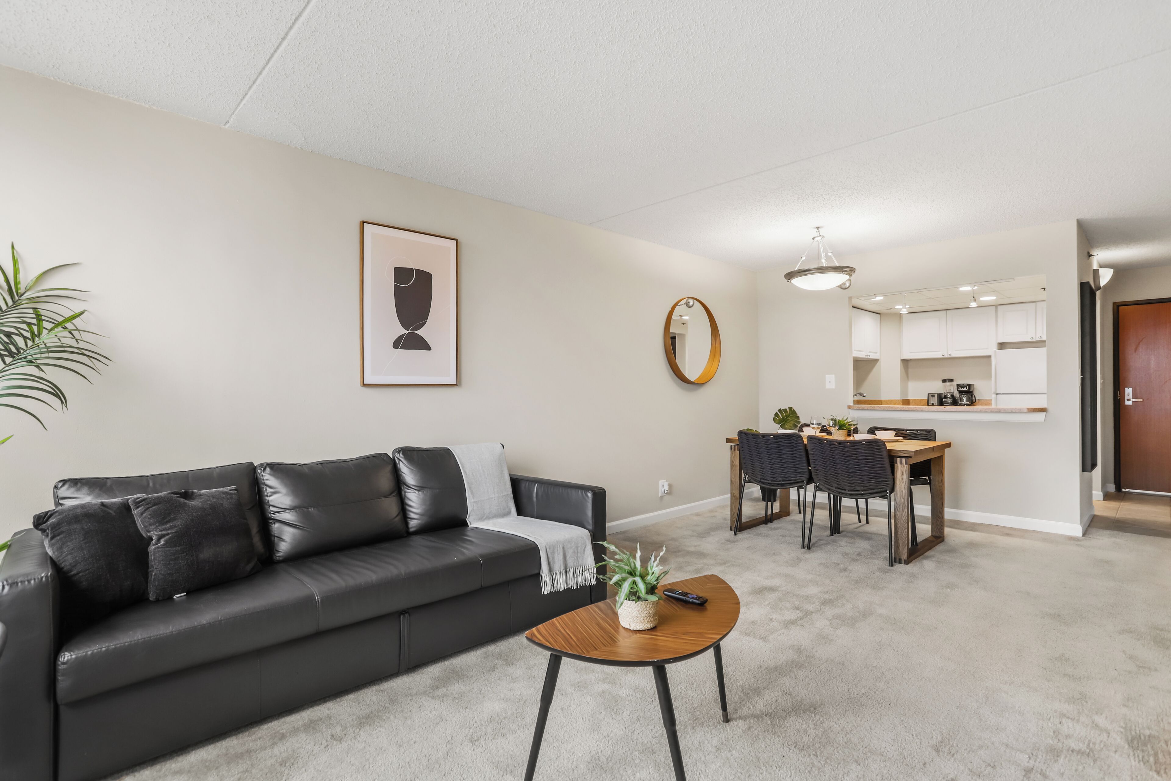Een woonkamer met een zwarte leren bank, een houten salontafel, een eethoek met tafel en stoelen, een keuken op de achtergrond en een hanglamp.