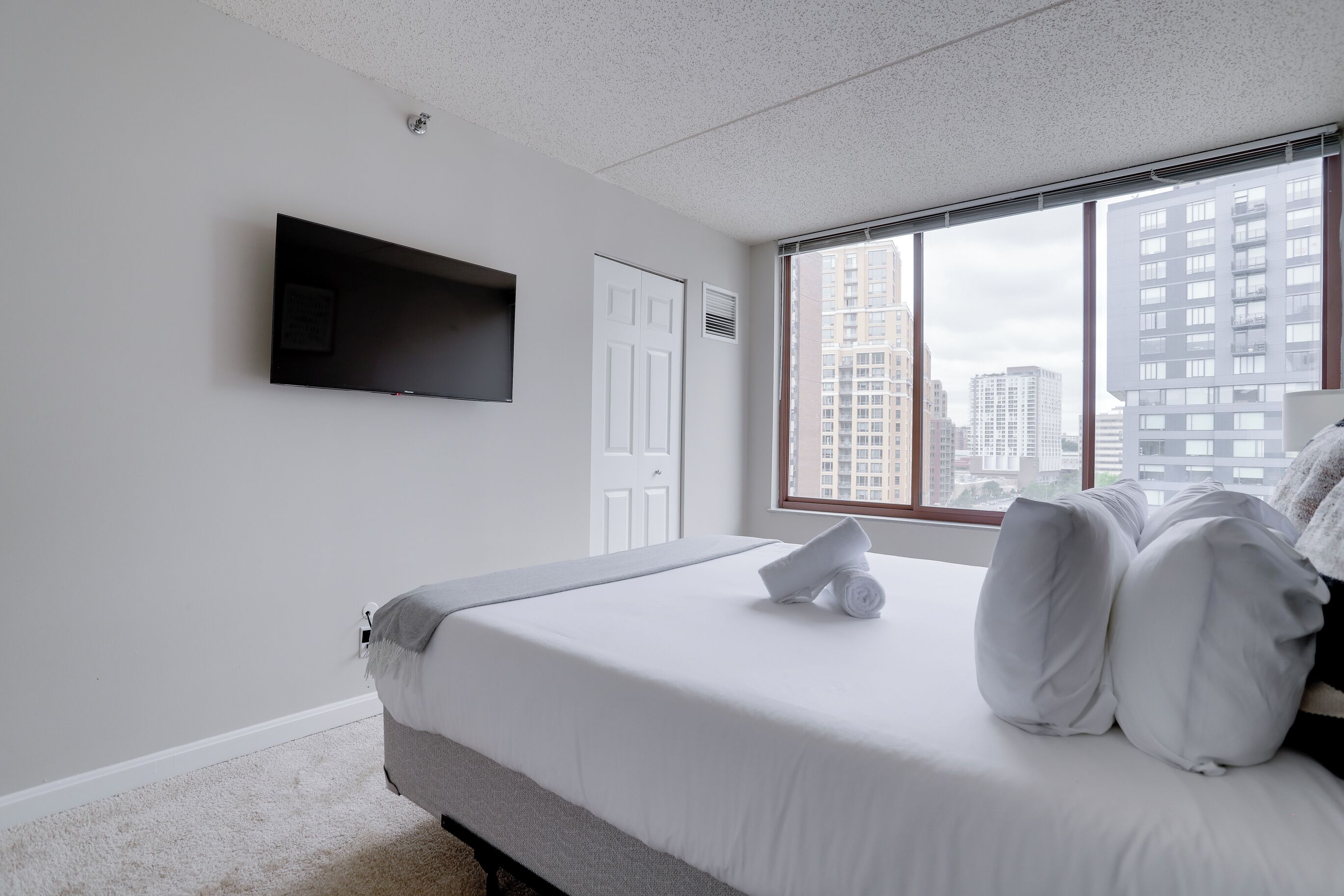 Une chambre d’hôtel avec un lit, un téléviseur à écran plat fixé au mur, une fenêtre donnant sur la ville et une porte.