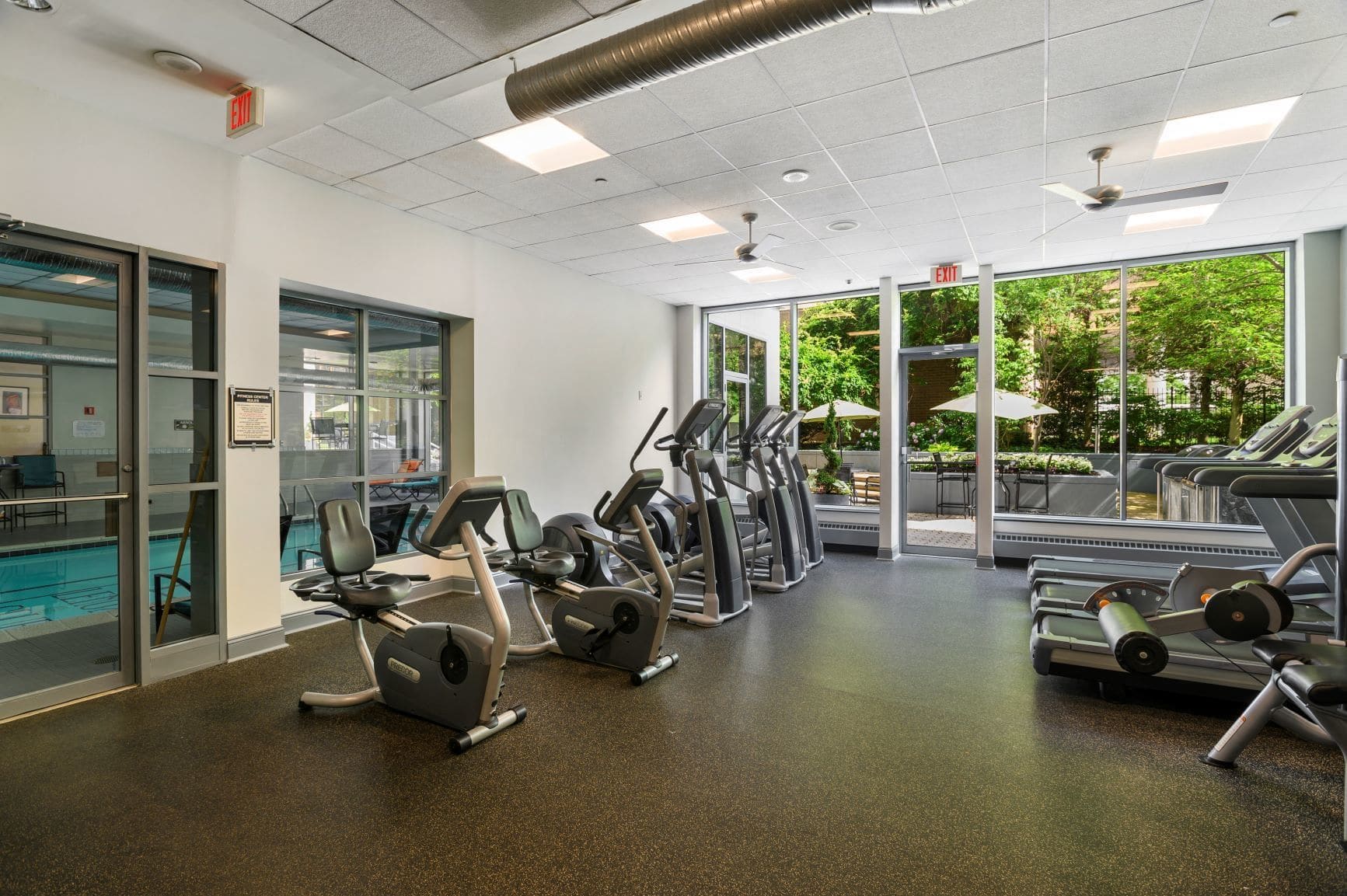 Une salle de sport équipée de tapis de course, de vélos elliptiques et d’appareils de musculation. Une porte vitrée donne accès à un espace piscine.