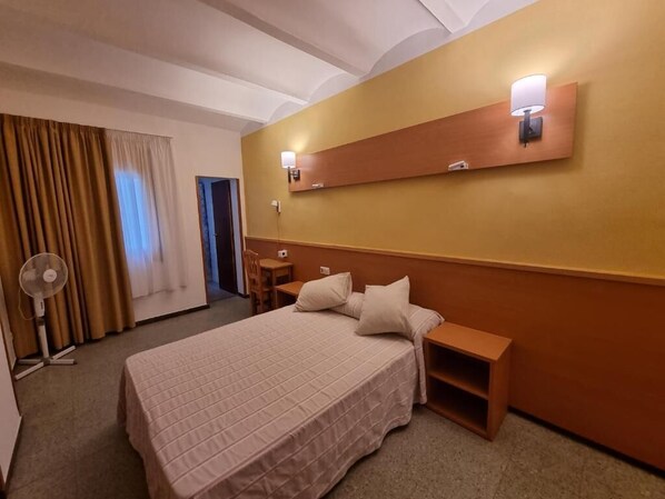A hotel room with a large bed, bedside tables, a desk, a chair, a standing fan, and a wall-mounted light fixture.