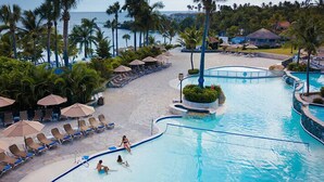 A resort pool area with lounge chairs and umbrellas, surrounded by palm trees and a view of the ocean. - Lifestyles (Puerto Plata)
