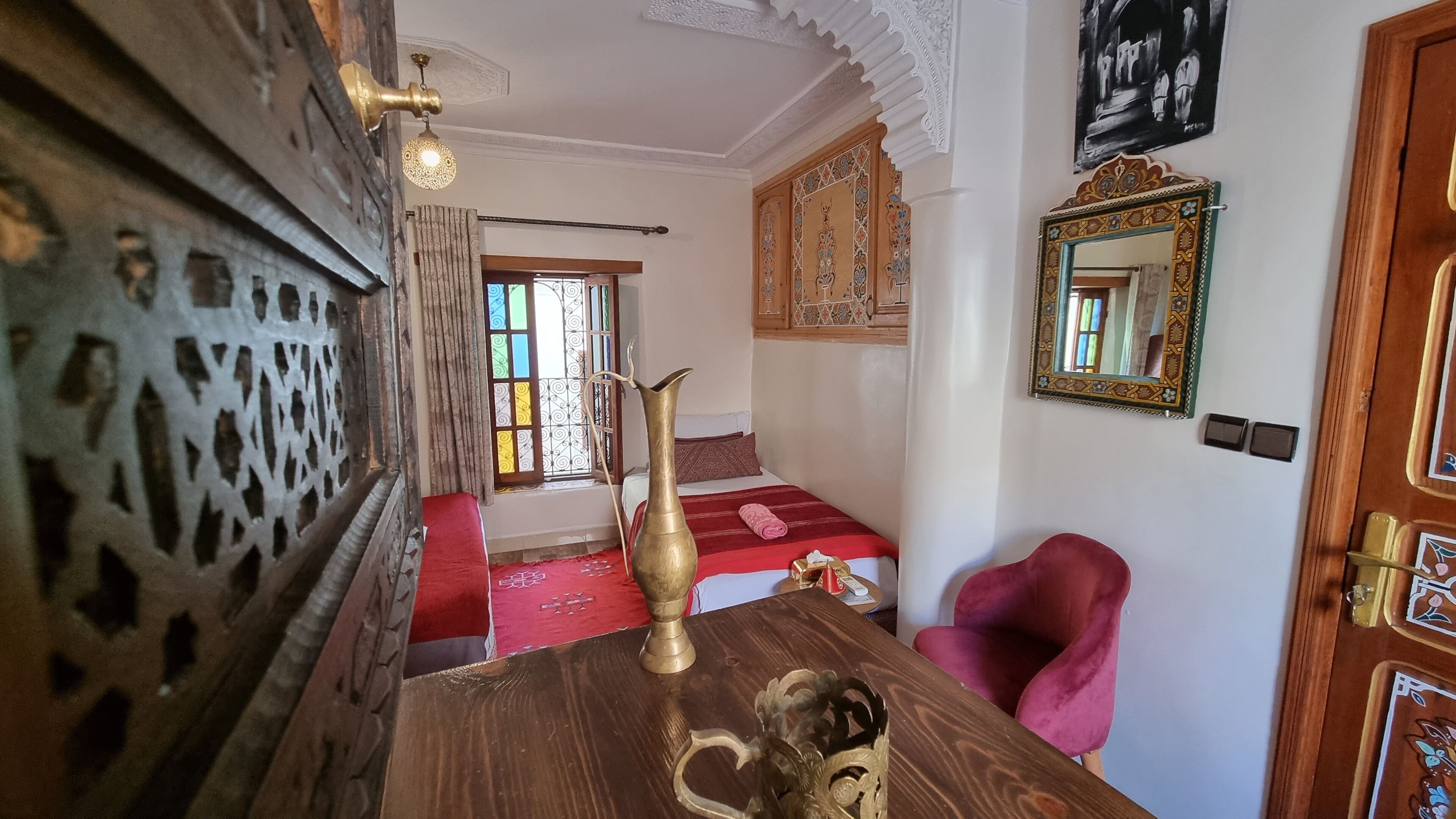 Una habitación con una mesa de madera, un jarro de latón, un sofá rojo, una silla rosa y una cama con colcha estampada.