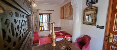 Una habitación con una mesa de madera, un jarro de latón, un sofá rojo, una silla rosa y una cama con colcha estampada.