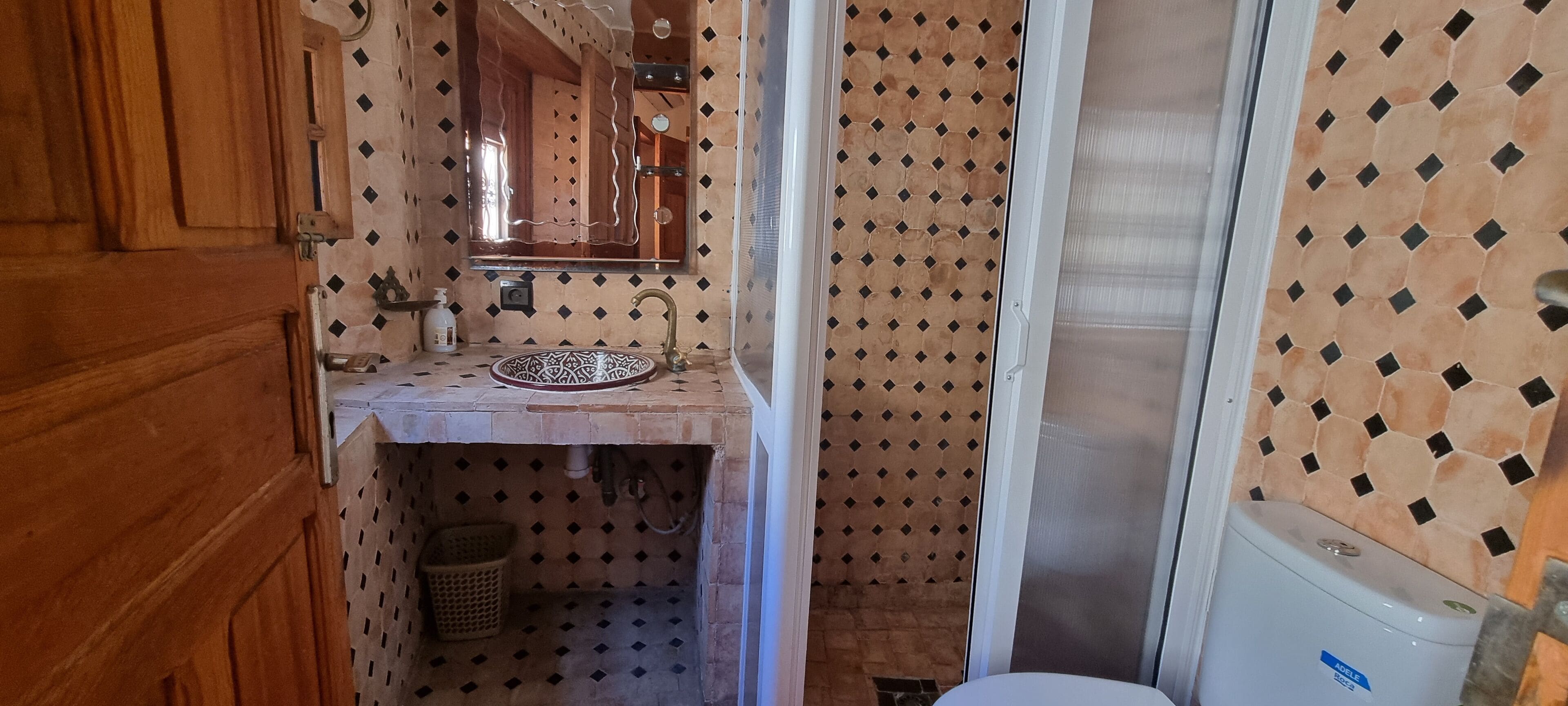 A bathroom with a wooden door, a sink with a patterned basin, a mirror, and a shower area with a tiled wall.