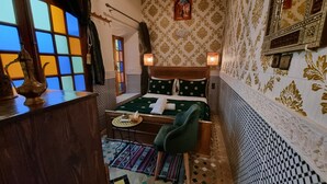 A room with a bed, a small table with a phone and a bowl, a chair, and a window with colorful panes. - Fes Touria Palace (Fes)