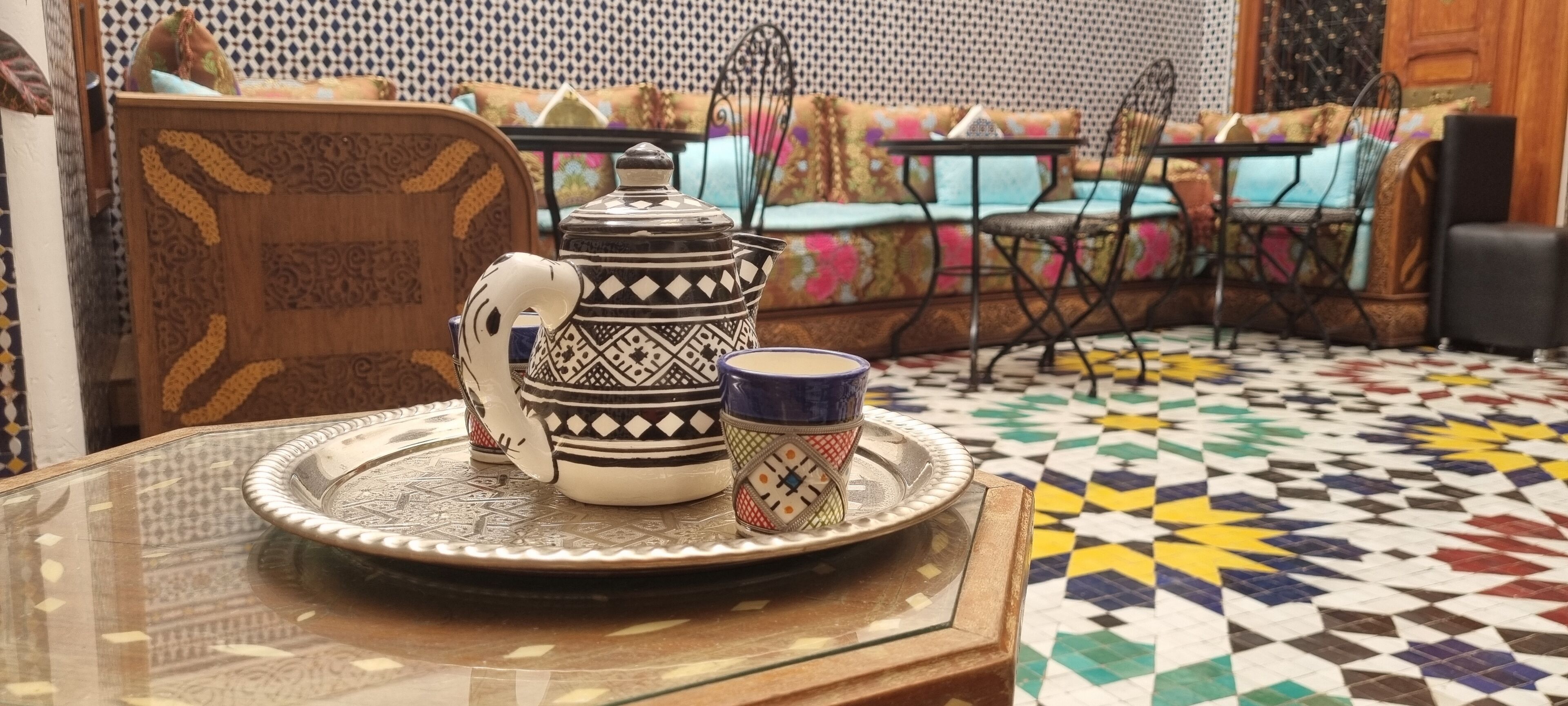 A traditional Moroccan tea set on a glass table with a patterned floor and colorful cushions in the background.