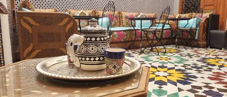 A traditional Moroccan tea set on a glass table with a patterned floor and colorful cushions in the background.