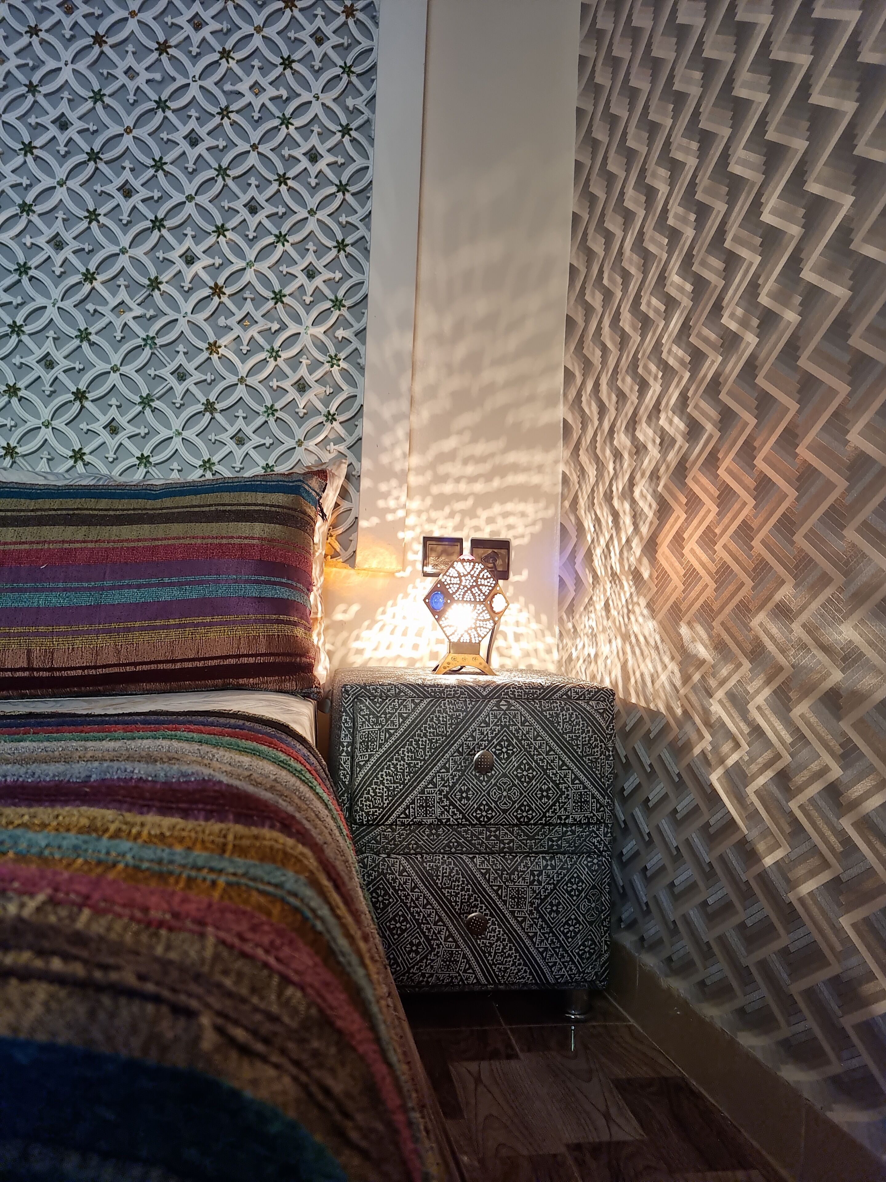 Una habitación con pared estampada, cama con manta a rayas coloridas y mesita de noche con lámpara decorada.