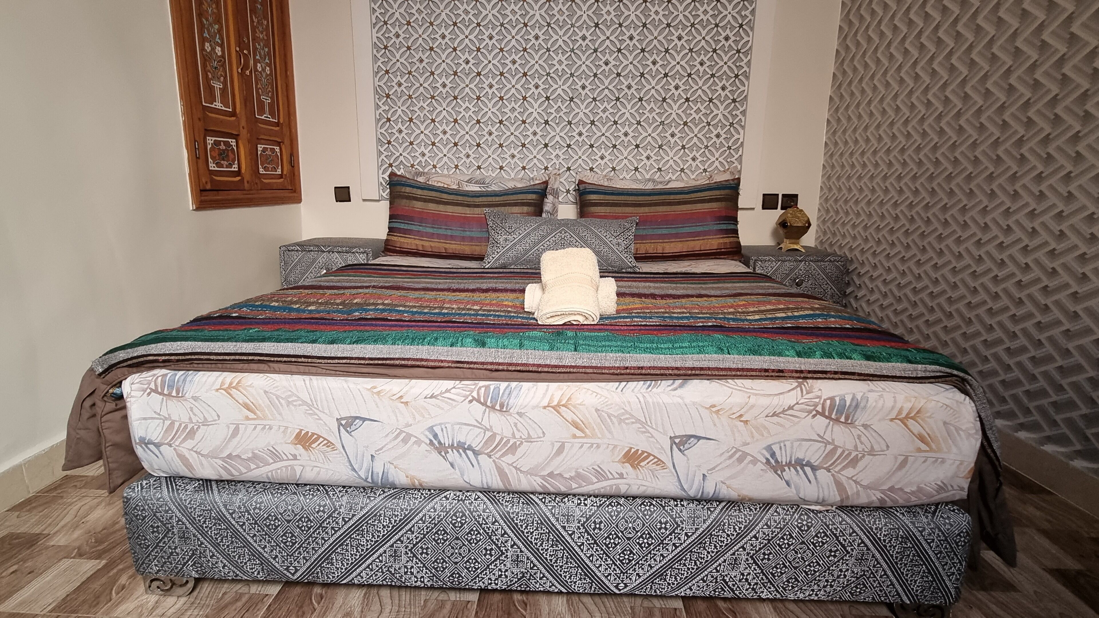 Una cama con una colcha estampada y almohadas, un panel decorativo en la pared y una puerta de madera con diseños intrincados.