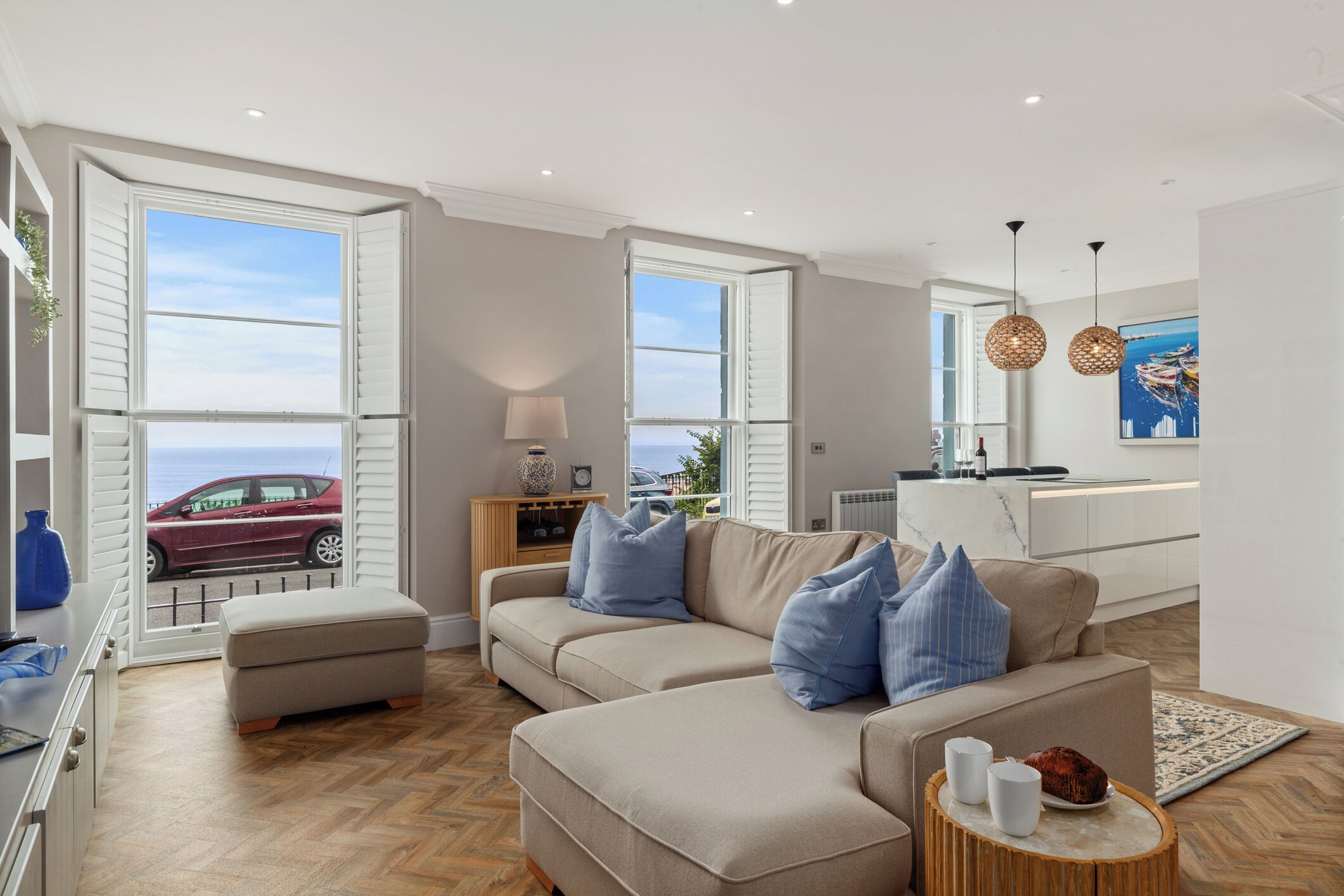 Un salon moderne avec un canapé beige, un pouf et une petite table d’appoint ronde en bois sur laquelle reposent deux tasses blanches. Une grande fenêtre donne sur une voiture rouge garée à l’extérieur.