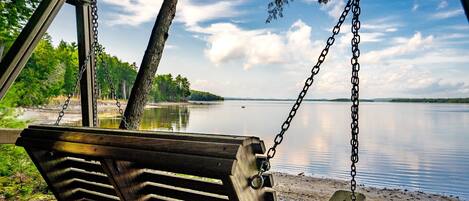 A wooden swing with metal chains, overlooking a calm lake with a forested shoreline.