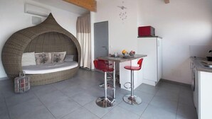Un espace de vie compact comprenant un canapé incurvé, une petite table à manger avec deux chaises rouges, un coin cuisine et une porte donnant sur une autre pièce.