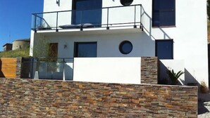Une maison moderne à deux étages, dotée d’une façade en pierre, de grandes fenêtres et d’un balcon.