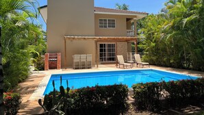 Une maison à deux étages avec une piscine, entourée d’une végétation luxuriante et de palmiers.