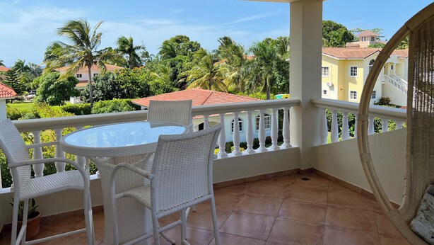 Un balcon avec une table et des chaises en osier peintes en blanc, un sol carrelé et une vue sur un quartier résidentiel, avec des palmiers et des maisons.