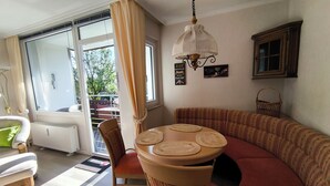 Una sala de estar acogedora con una mesa redonda, un sofá y una silla. Hay una ventana con cortinas, un radiador y una lámpara colgante.