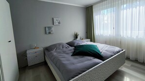 Un dormitorio moderno con cama, mesitas de noche y una ventana con cortinas traslúcidas.
