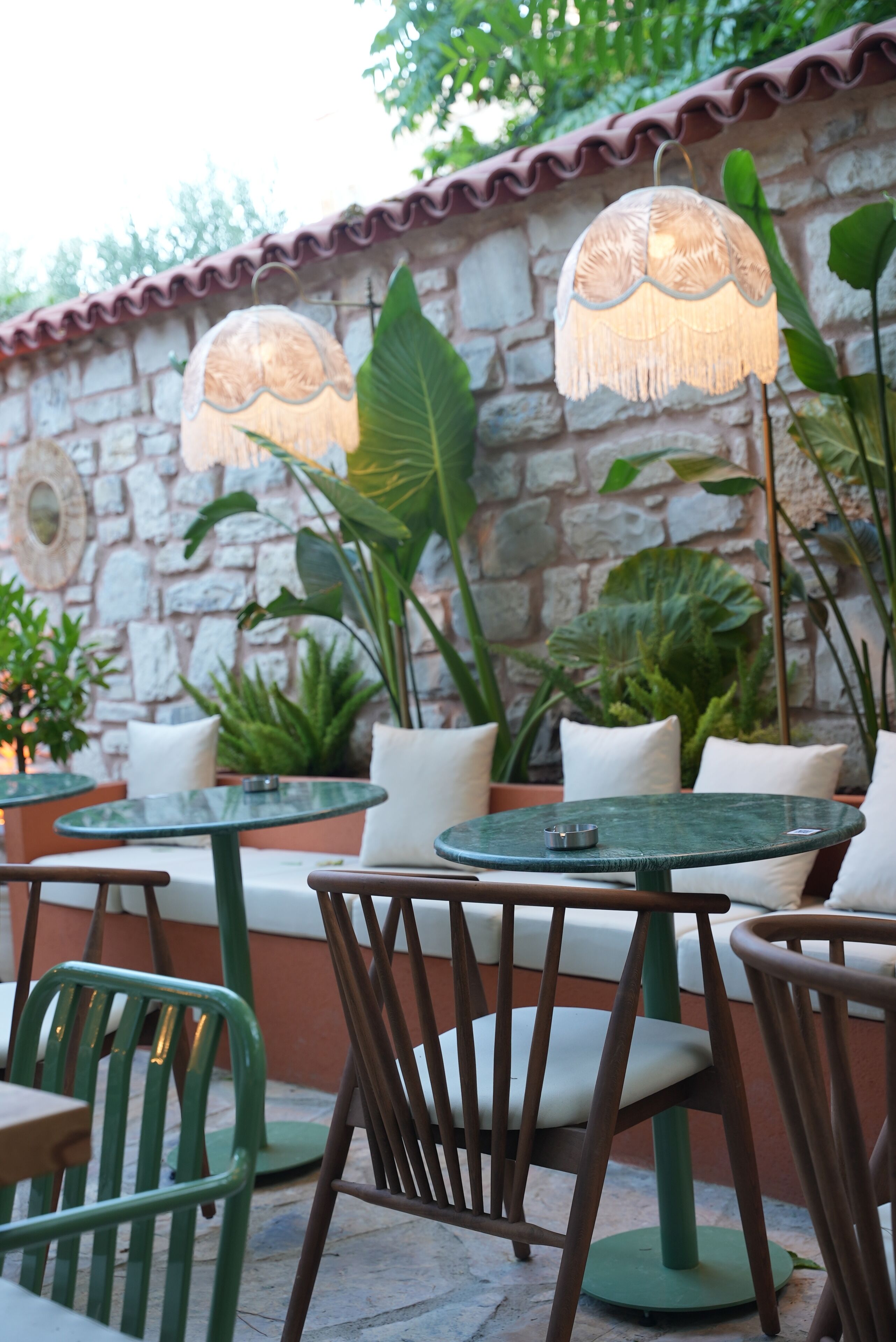 Terraza al aire libre con mesas y sillas, rodeada de plantas en macetas y un muro de piedra.