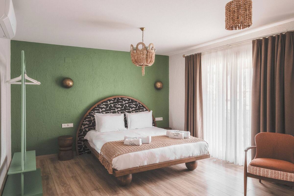 A neatly made bed with a patterned headboard, a hanging woven light fixture, and a view of a window with curtains.