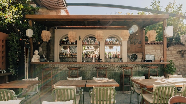 An outdoor bar area with wooden tables and chairs, a stone wall, and a bar counter with bottles and glasses. - Pietra Social House (Urla)