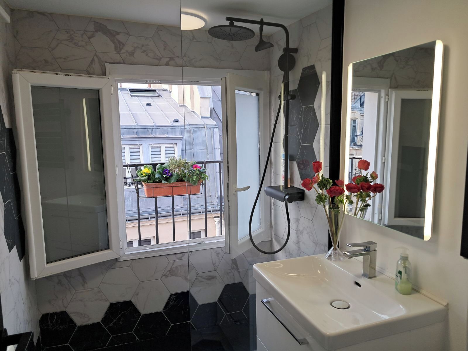 A bathroom with a white sink, a mirror, a shower, and a window with a balcony view.