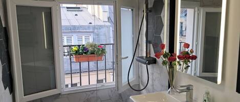 A bathroom with a white sink, a mirror, a shower, and a window with a balcony view.