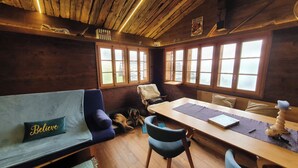 Ein gemütliches Blockhausschlafzimmer mit Holzbalkendecke, einer Couch mit Kissen mit dem Aufdruck 'Believe', einem Esstisch und einem Fenster mit Buntglas.