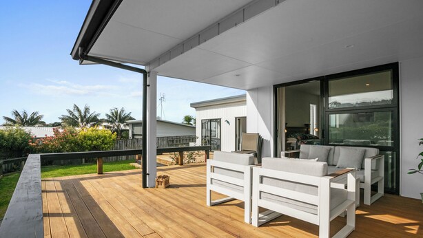 A covered outdoor area with wooden flooring, white furniture, and large windows offering views of the surrounding landscape. - Beachy Serenity - Waihi Beach Holiday Home (Waihi Beach)