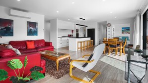 A modern living room with a red sofa, a glass coffee table, and a dining area with wooden chairs and a table. - Beachy Serenity - Waihi Beach Holiday Home (Waihi Beach)