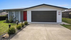 A single-story house with a garage, a red door, and a small front yard with plants and a concrete driveway. - Beachy Serenity - Waihi Beach Holiday Home (Waihi Beach)