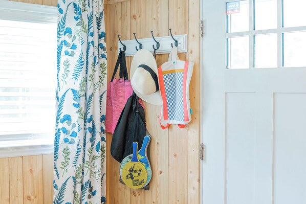 A room with wooden walls, a window with blinds, a curtain with a blue and white pattern, and various items hanging on a wall-mounted rack, including hats, bags, and a paddleboard. - NEW-The Beach Haus- Luxe Cottage, professionally designed w/ WiFi, linens incl. (Supply)