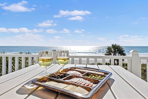 Una bandeja de comida y dos copas de vino blanco sobre una mesa de madera con vista al océano.