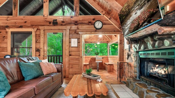 A cozy cabin interior with a wooden sofa, a dining table, a fireplace, and a large window overlooking greenery. - Romantic private cabin near Pigeon Forge - Hot Tub, Fireplace, Views!!! (Pigeon Forge)