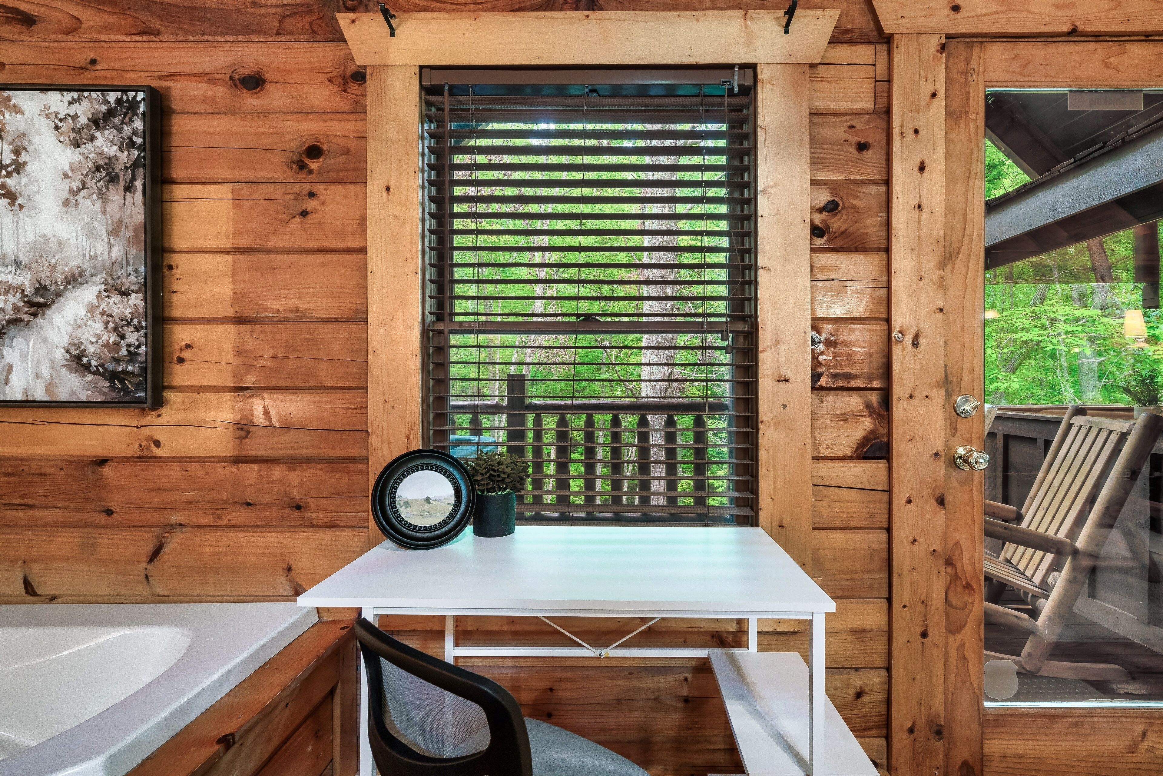 A wooden cabin interior with a white desk, a black chair, a window with blinds, and a view of greenery outside.