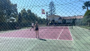 Un court de tennis entouré d’une clôture en treillis métallique, bordé d’arbres et avec un bâtiment visible au loin.