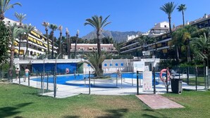 Un espace piscine avec une piscine entourée d’une clôture, des palmiers et des bâtiments en arrière-plan.