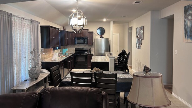 A modern kitchen with dark wood cabinets, a stainless steel refrigerator, and a black stove. There is a dining area with a table and chairs, and a living room with a leather sofa and a lamp.