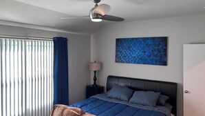 A bedroom with a bed, a ceiling fan, a window with curtains, a nightstand with a lamp, and a wall art piece.