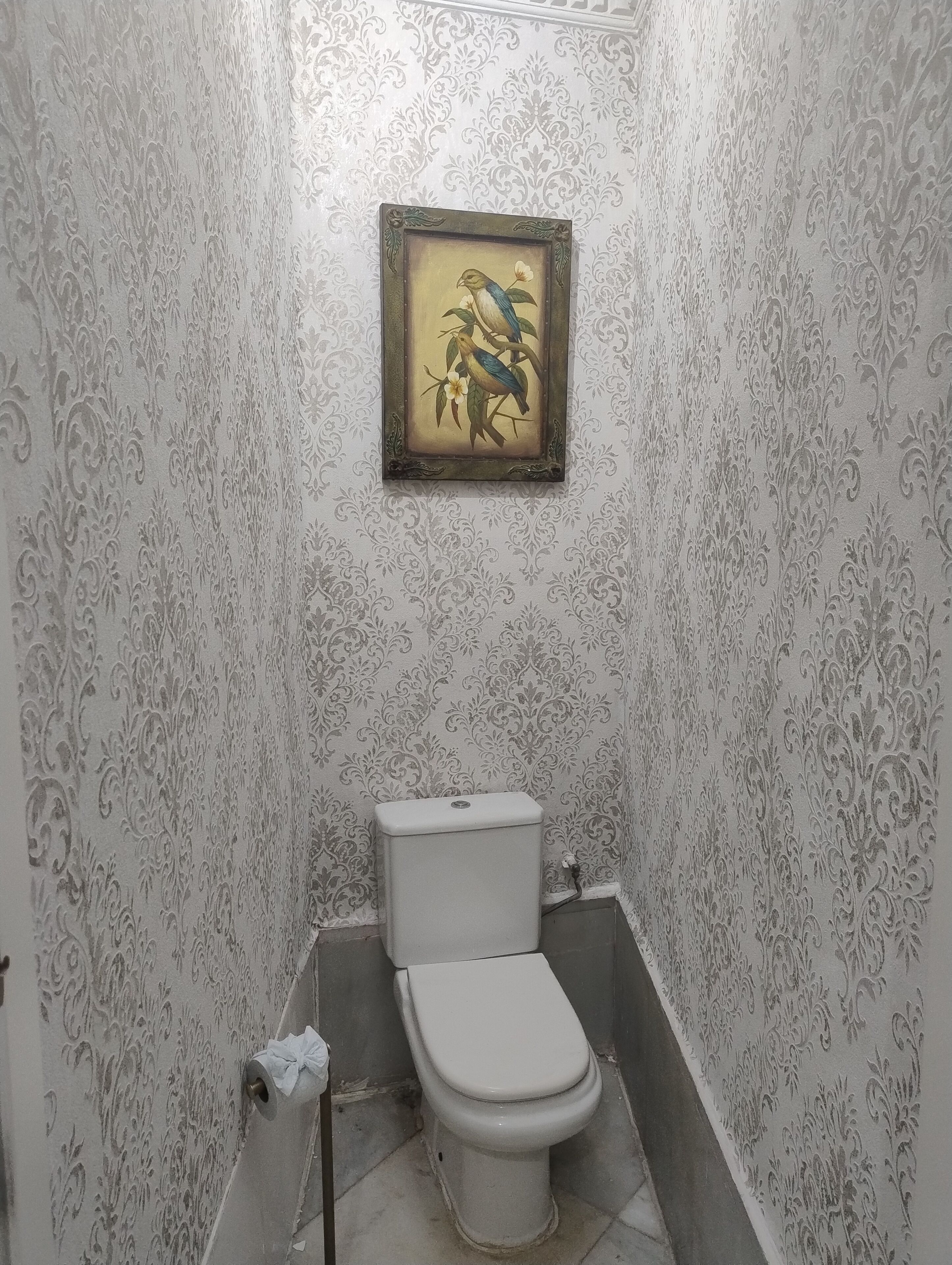 A small bathroom with a toilet, a framed picture on the wall, and patterned wallpaper.
