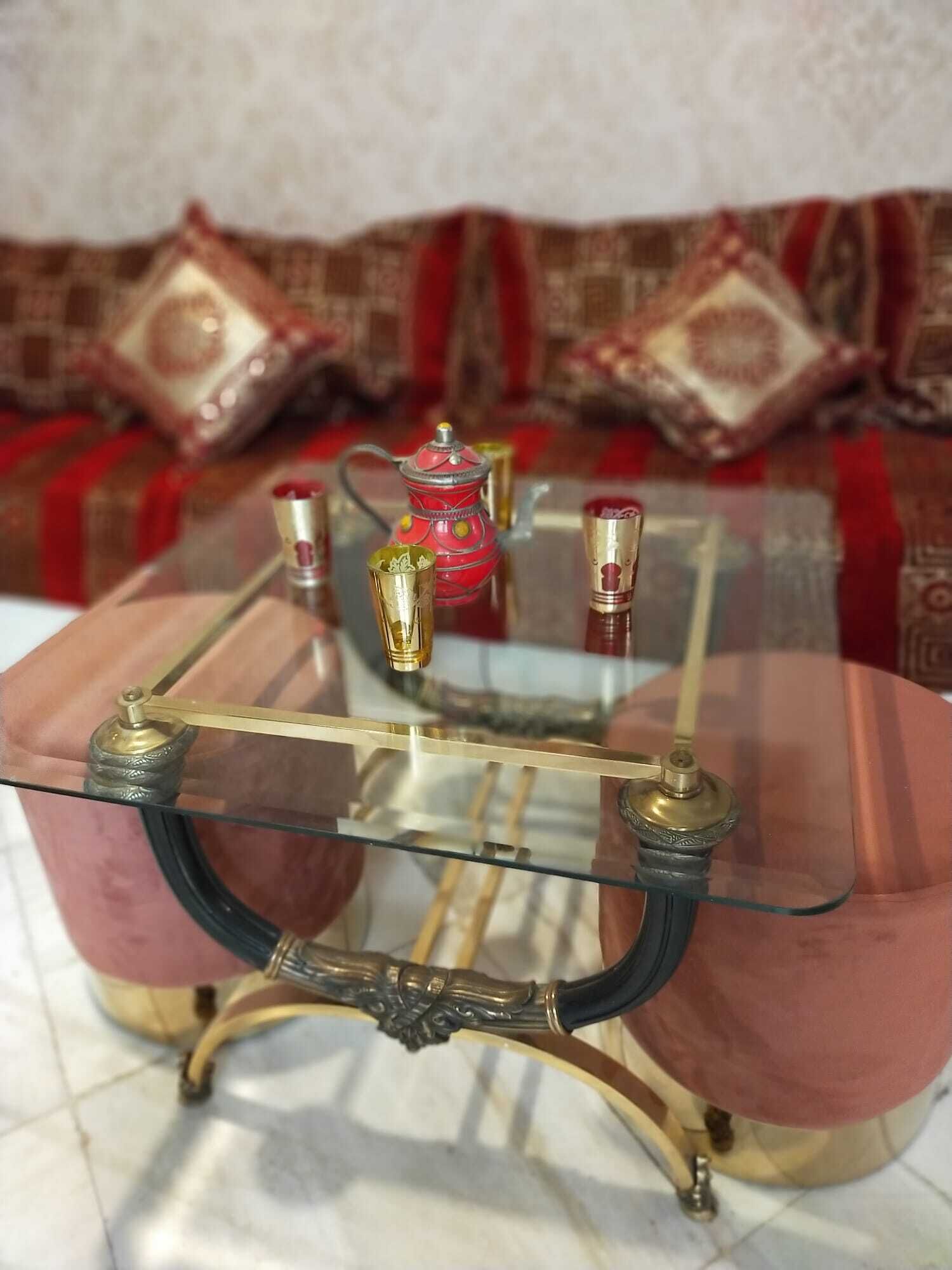 Una mesa con cubierta de vidrio y patas de metal ornamentadas, una tetera roja y dos jarrones pequeños sobre un sofá estampado.
